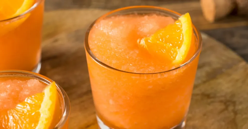 Three glasses of orange slushi garnished with fresh orange wedges on a wooden surface