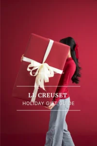 Person holding oversized red gift box with bow on red background, representing Le Creuset holiday gift guide.